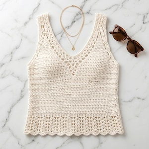 May include: A cream-colored sleeveless crochet top featuring a V-neckline with an intricate openwork pattern, a solid midsection, and a scalloped hem. Accompanying the top is a delicate gold-toned layered necklace and brown tortoiseshell sunglasses.