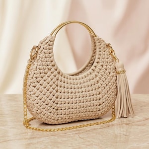 Può includere: Borsa a mano all'uncinetto beige con manico e tracolla a catena color oro. La borsa presenta una forma arrotondata e un dettaglio a nappa. La borsa è realizzata in materiale tessuto.