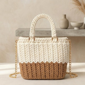 May include: A cream and brown crochet handbag with a gold chain strap. The bag has a cream-coloured handle and a two-tone design. The bag is a small tote style, suitable for everyday use.