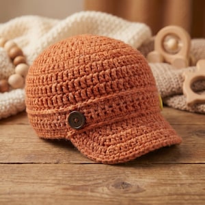 Può includere: Un cappello arancione all'uncinetto con una piccola visiera e un bottone marrone. Il cappello è realizzato con filato strutturato e sembra fatto a mano. Il cappello è su una superficie di legno.