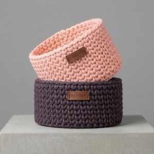 May include: Two hand-crocheted baskets, one stacked on top of the other. The top basket is light pink, and the bottom basket is dark purple. Each basket has a brown leather tag that reads "WHISPER".