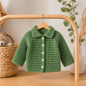 May include: A green crochet jacket with a collar and button closure, hanging on a wooden hanger. The jacket is made of a textured yarn and has long sleeves. The buttons are white. The jacket is displayed on a wooden rack.