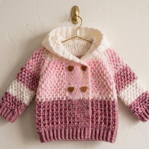 May include: A handmade, pink and white crochet baby jacket with a hood. The jacket features a double-breasted design with wooden buttons. The sleeves have white stripes. The jacket is hanging on a gold hanger.