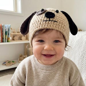 May include: A tan crochet dog hat with black and brown accents. The hat features a dog face design with floppy ears and a black nose. The child is wearing a tan sweater.