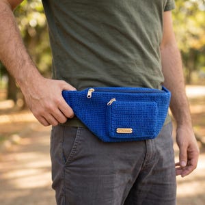 May include: A bright blue crochet fanny pack featuring a main zippered compartment and a smaller front zippered pocket. A rectangular light-colored wooden tag with the text Nissa is attached to the lower right corner of the front pocket.