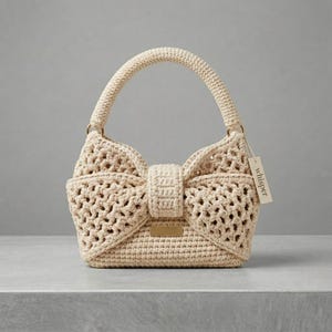 Op de afbeelding: Een crèmekleurige gehaakte handtas met een strikvormig ontwerp. De tas heeft een gestructureerde handgreep en een klepsluiting. Een klein label met het woord "whisper" is bevestigd. De tas is gemaakt van een getextureerd, open geweven materiaal.