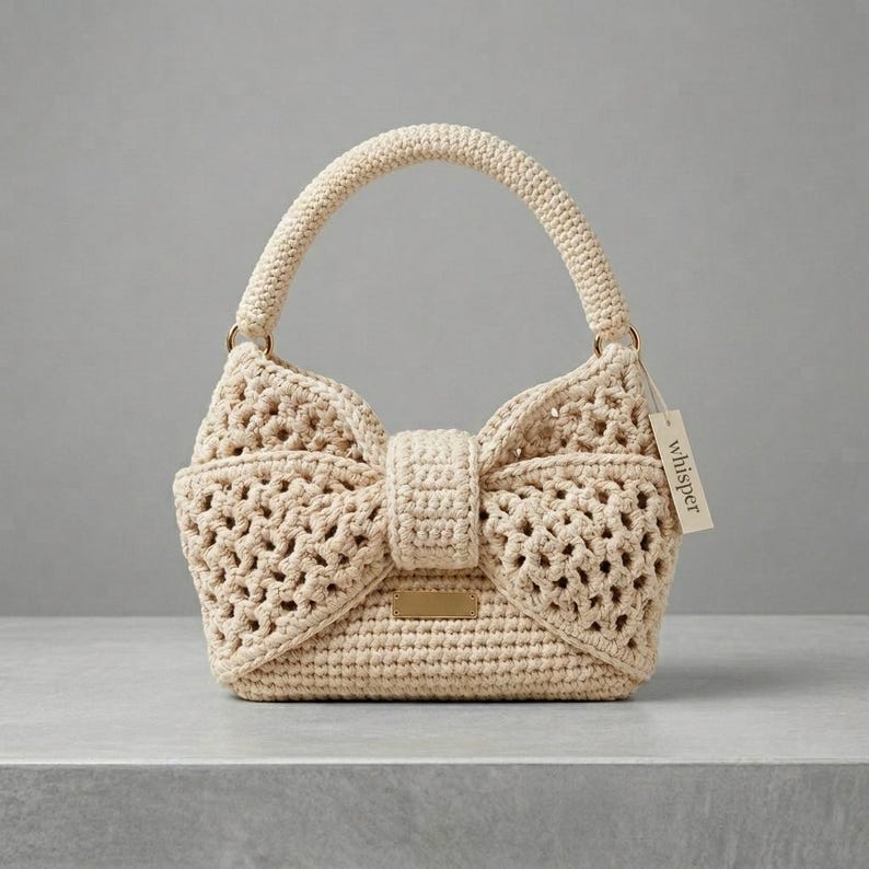 Peut inclure: Un sac &agrave; main en crochet de couleur cr&egrave;me avec un design en forme de n&oelig;ud papillon. Le sac est dot&eacute; d'une poign&eacute;e structur&eacute;e et d'une fermeture &agrave; rabat. Une petite &eacute;tiquette avec le mot "whisper" est attach&eacute;e. Le sac est en mat&eacute;riau textur&eacute; et ajour&eacute;.