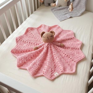 Könnte beinhalten: Eine handgefertigte rosa Baby-Häkeldecke mit Sternmuster und einem kleinen, braunen Bärenkopf und Armen. Die Decke liegt auf einem weißen Bettlaken.