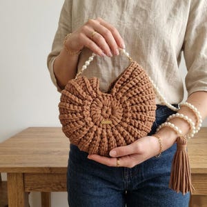 Peut inclure: Un sac à main marron crocheté avec un design unique en forme de coquillage. Le sac est doté d'une bandoulière en perles et d'un pompon assorti. La personne qui le tient porte des bracelets et des bagues dorés. Le sac a une petite étiquette dorée.