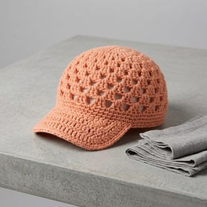 Puede incluir: Gorra de ganchillo color melocotón con visera. La gorra tiene un diseño abierto y aireado, y está hecha de un hilo suave y texturizado. La gorra está sobre una superficie gris junto a un paño gris doblado.
