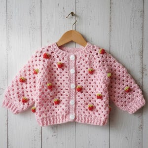 Peut inclure: Un cardigan rose clair en crochet avec un motif de fraises. Le pull à boutons présente des appliqués de fraises rouges et vertes et est présenté sur un cintre en bois sur un fond en bois blanc.