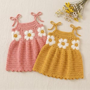 May include: Two sleeveless crochet dresses, one in dusty pink and one in mustard yellow, each featuring three white daisy motifs with yellow centres across the chest, tie-up shoulder straps, and a scalloped hemline with a textured knit pattern.