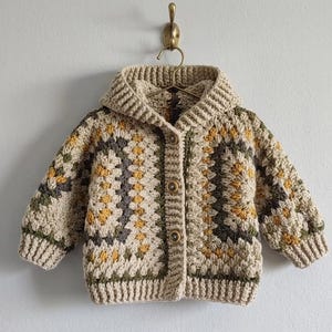 May include: A beige, hooded crochet cardigan with a button-up front. The sweater features a geometric pattern in shades of olive green, mustard yellow, and grey. The garment is hanging on a brass hook against a white wall.