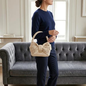 Peut inclure: Un sac &agrave; main beige en crochet avec un motif en forme de n&oelig;ud papillon. Le sac a une forme structur&eacute;e avec une poign&eacute;e et une &eacute;tiquette attach&eacute;e. Le sac est tenu par une personne portant un pull et un pantalon bleu marine.