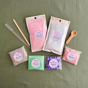 Peut inclure: Ensemble de matériaux de jeu sensoriels, dont une poudre rose "Mermaid Scale Powder", des perles irisées et des "Magic Mermaid Eggs" violets. Une cuillère en bois, une pipette et une baguette sont également incluses. Les produits sont emballés dans des sachets transparents.
