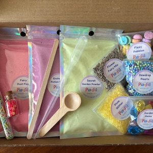 May include: A sensory play kit in a cardboard box containing three large iridescent pouches of pink, purple, and green powder, a clear plastic pipette, a wooden scoop, a test tube of sprinkles, and small bags of pebbles, pearls, glitter, and gems.