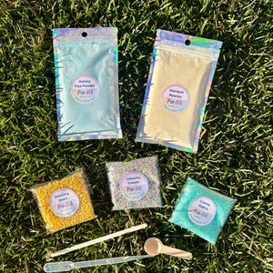 May include: A selection of craft supplies, including powders and beads, in clear bags, arranged on grass. The bags are labelled "Galaxy Fizz Powder," "Stardust Reactor," "Shooting Stars," "Colorful Comets," and "Cosmic Debris." Wooden scoops and droppers are also included in the image.