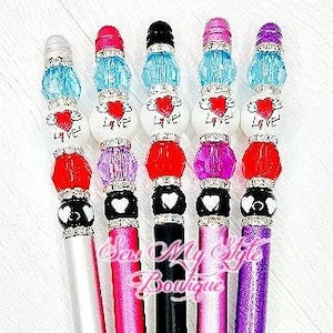 May include: A collection of decorative pens with jewel-toned accents. Each pen features a different color scheme, including silver, pink, and black barrels, topped with beads and crystals. The pens have a heart and the word "Love" on them.