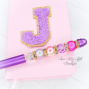 May include: A pink notebook with a purple letter J on the front. The notebook is closed and has a purple pen with pink and silver beads attached to it. The pen has the text "Sew My Style Boutique" on it.