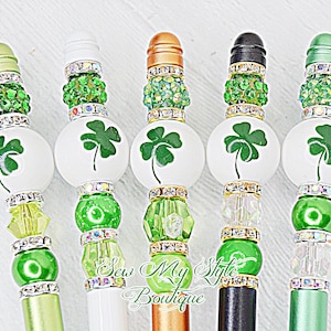 May include: Five decorative pens with green shamrock designs and crystal embellishments. The pens have different colored tops and bases, including green, white, black, gold, and silver.