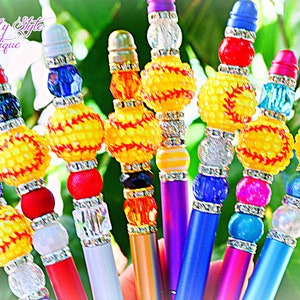 May include: A set of ten colorful pens decorated with beads and a softball charm. The pens are in various colors, including purple, red, blue, and green. The softball charm is yellow with red stitching.