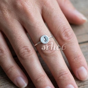 Puede incluir: Un anillo de tono plateado que presenta un pequeño dije circular y plano. El centro del disco está estampado con la letra mayúscula J en una fuente oscura y hundida. La banda del anillo es delgada y metálica, rodeando el dedo con sencillez.