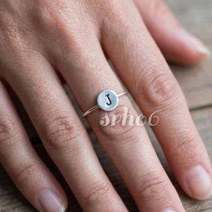 May include: A silver-toned ring featuring a thin band and a small, circular flat disc centerpiece. The disc is stamped with the uppercase letter J in a dark, recessed font. The metal has a matte, brushed finish throughout the entire piece.