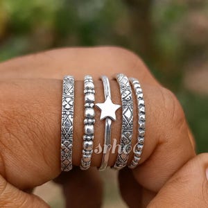 May include: A set of five silver-toned rings worn together. The rings feature varied textures including a geometric pattern, a beaded design, a smooth band with a central five-pointed star, an intricate etched pattern, and a small textured bead band.