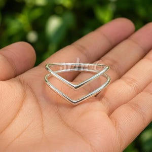 May include: A polished silver ring featuring a double chevron or V-shaped design. The band consists of two parallel, smooth silver wires that meet at a central point, creating a geometric, open-style silhouette with a bright, reflective finish.
