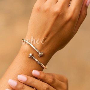 May include: A silver-toned metal cuff bracelet shaped like a bent nail, featuring a smooth, polished finish and a textured head design at each end. The bracelet wraps around the wrist with an open-ended, bypass-style construction.