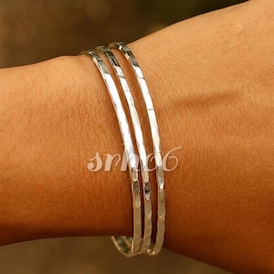 May include: Three thin, silver-coloured metal bangles with a hammered, textured surface. Each bangle features a reflective finish that catches the light, creating a series of small, indented facets along the entire circumference of the circular bands.