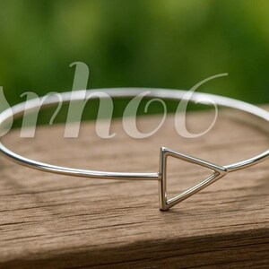 May include: A thin, polished silver-tone metal bangle bracelet featuring a single open triangle shape as a focal point. The bracelet has a smooth, rounded wire band that connects to the geometric triangle element on one side of the circular frame.