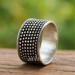 Puede incluir: Un anillo de banda ancha de plata con una superficie texturizada cubierta por un patrón repetitivo de pequeños puntos metálicos en relieve. El exterior del anillo tiene un acabado oscuro y oxidado entre las esferas de plata pulida, con interior liso.