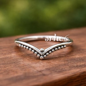 Puede incluir: Un anillo de plata en forma de chevron con una delicada textura de cuentas a lo largo del borde exterior de la banda en V. El metal tiene un acabado pulido con acentos oscuros entre las pequeñas esferas, y el texto srho6 es visible en blanco.