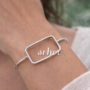 May include: A silver-toned metal bangle bracelet featuring a central rectangular frame design. The bracelet has a smooth, polished finish and a thin, open-ended wire construction that wraps around the wrist. The text "srhot" is visible in white.