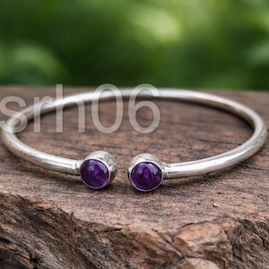 May include: A silver cuff bracelet featuring two round, polished purple amethyst gemstones set at each open end. The metal band has a smooth, reflective finish, and the two stones are identical in size and deep violet color, creating a balanced look.