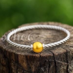 May include: A silver-toned twisted cable bangle bracelet featuring a single, smooth, spherical yellow-gold colored bead at the center, flanked by two small silver-toned cylindrical accents on either side of the central bead.