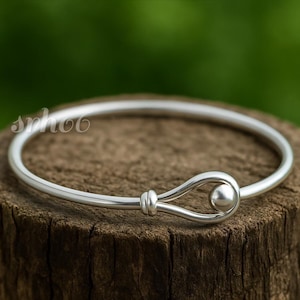 May include: A polished silver bangle bracelet featuring a smooth, circular band that terminates in a hook-and-eye style clasp. The clasp consists of a small, rounded silver bead secured within a looped wire end, creating a clean, continuous look.