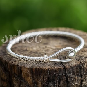 Könnte beinhalten: Ein silberfarbener Armreif mit einer gedrehten, seilartigen Textur entlang des Bandes. Das Armband endet in einem Hakenverschluss mit einer glatten, abgerundeten Schlaufe und einem kleinen kugelförmigen Detail. Der Text "sphoo" ist auf der linken Seite zu sehen.