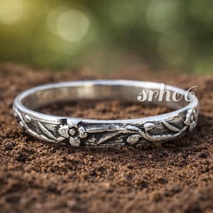 Könnte beinhalten: Ein Silberring mit einem erhabenen, geprägten Blumen- und Rankenmuster rund um das gesamte Band. Das Metall hat ein abgedunkeltes, oxidiertes Finish in den vertieften Bereichen, um die botanischen Details hervorzuheben. Text srho6 ist sichtbar.