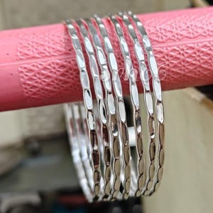 Puede incluir: Un conjunto de seis brazaletes plateados, cada uno con una textura martillada distintiva en la superficie exterior. Los brazaletes están dispuestos en una fila paralela, mostrando un acabado metálico reflectante y un perfil circular delgado.