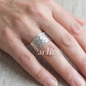 Può includere: Un ampio anello in argento composto da cinque fasce individuali, sottili e lisce, impilate insieme per creare un unico pezzo coeso. Il metallo ha una finitura lucida e riflettente che cattura la luce, mostrando un design uniforme e pulito.