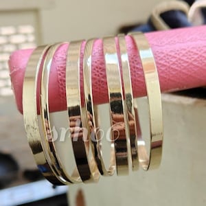 Puede incluir: Un conjunto de seis brazaletes de metal en tono dorado pulido con una superficie lisa y plana. Las pulseras están dispuestas en fila, mostrando un acabado metálico reflectante y un diseño circular simple y uniforme de ancho constante.