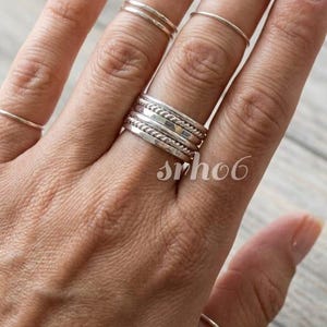 Könnte beinhalten: Ein Set aus silberfarbenen Ringen, die an mehreren Fingern getragen werden. Der Mittelfinger zeigt einen Stapel aus vier verschiedenen Ringen, darunter glatte, gedrehte und strukturierte Designs. Weitere dünne, schlichte Ringe an anderen Fingern.