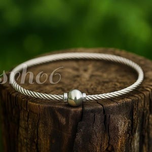 Può includere: Un bracciale a cavo color argento caratterizzato da una fascia in filo metallico intrecciato e una perla centrale liscia e sferica. Il bracciale ha una finitura lucida e una forma circolare, con il testo "scho6" visibile in bianco a sinistra.