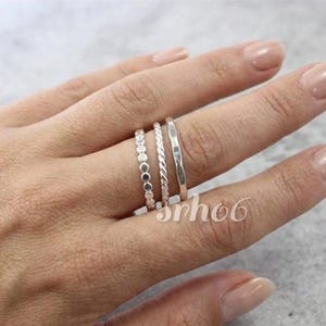 Könnte beinhalten: Ein Set aus drei silberfarbenen Ringen an einem Finger. Der erste Ring besteht aus einer Reihe kleiner, verbundener runder Perlen. Der zweite Ring hat eine gedrehte, seilartige Textur. Der dritte Ring ist ein glattes Band mit Hammerschlag.