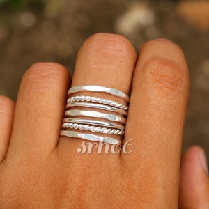Può includere: Una pila di sei anelli in argento indossati su un dito. Il set comprende tre fasce lisce con texture martellata e due fasce a corda intrecciata, tutte con una finitura metallica lucida. Il testo "srho6" è visibile sugli anelli inferiori.