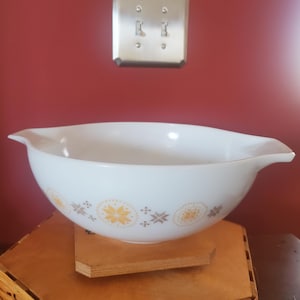 May include: White Pyrex mixing bowl with a decorative pattern of yellow and brown starburst and floral designs. The bowl has two handles and is sitting on a wooden surface. A red wall and light switches are in the background.