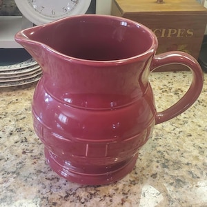 May include: A burgundy ceramic pitcher with a handle and spout. The pitcher has a textured, banded design around the base and body. The pitcher is sitting on a speckled countertop.