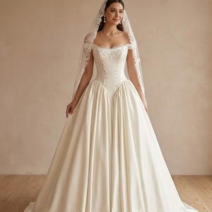 Op de afbeelding: Een gebroken witte, vloerlange japon met een off-shoulder halslijn en een aansluitend lijfje met gedetailleerd wit borduurwerk. De rok is vol en volumineus met een gladde satijnen afwerking. Een lange, transparante sluier met kanten rand.